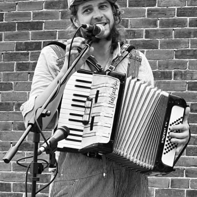 Black and white picture of Benj Rowland playing an accordian. He is standing in front of a brick wall
