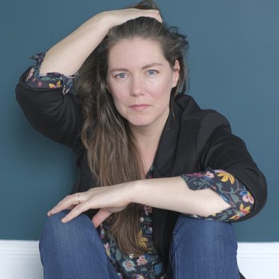 Catherine MacLellan leaning against a teal wall with one arm on her head and another arm crossed over her knees