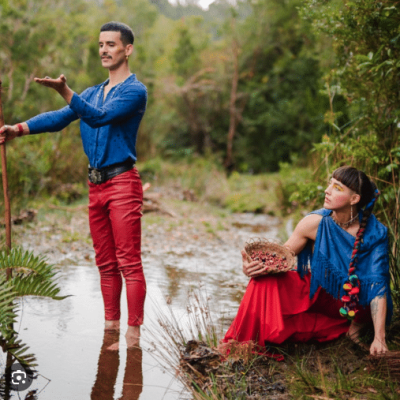 Chola Y Gitano duet sitting creekside in red and blue outfits