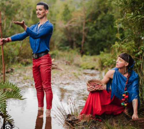 Chola Y Gitano duet sitting creekside in red and blue outfits