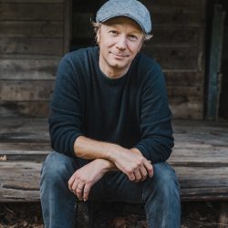 Daryl Chonka sitting on step, wearing a blue hat, sweater and jeans, sitting on the deck edge of a cabin