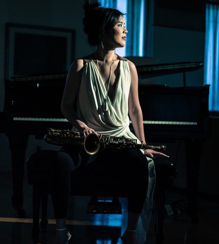 Stephanie Chou sitting on a piano bench in front of a piano, with a saxophone in her lap. She is highlighted by light in a darkened room that contains turquoise curtains. Credit photo Emra Islek.