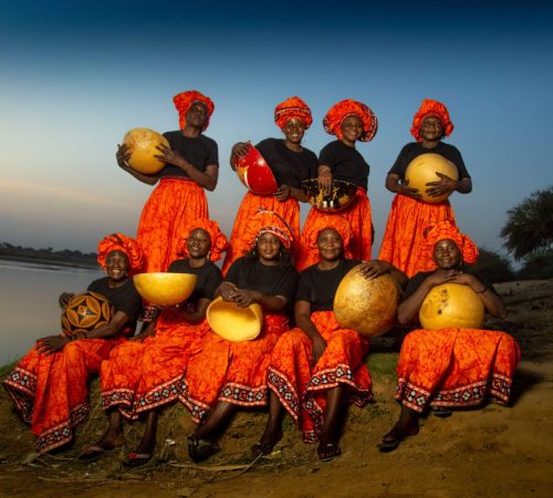 Ten women clad in orange dresses holding wooden bowls. They all are sitting beside a riverbank