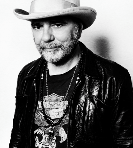Black and white photo of Daniel Lanois. He is wearing a black leather jacket, with a Harley Davidson shirt underneath and chain type necklaces. He is also wearing a white felt cowboy hat adorned with a ribbon around the hat.
