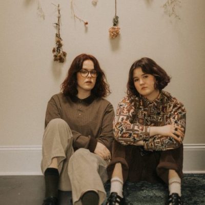 Moira and Claire sitting on floor with beige wall behind them that contains dried flowers affixed to the wall